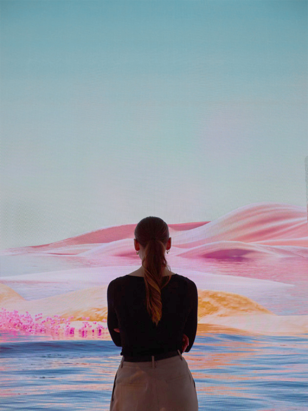 A woman with a ponytail stands with her back to the viewer, engrossed in a projected display of a vivid, dreamlike desert landscape