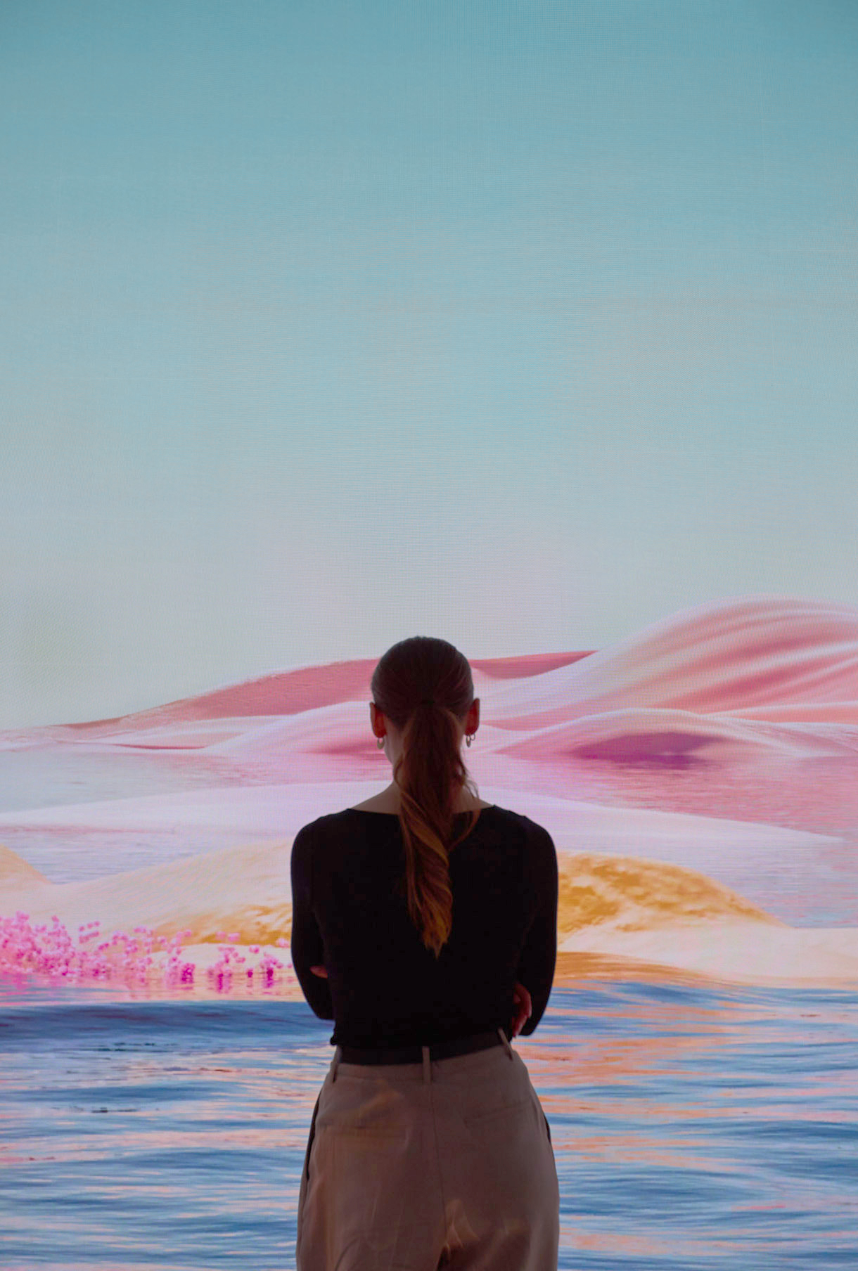 A woman with a ponytail stands with her back to the viewer, engrossed in a projected display of a vivid, dreamlike desert landscape