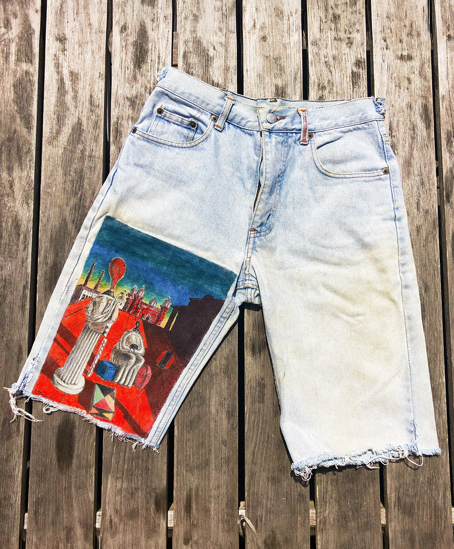 Light blue denim shorts placed on a wooden floor, with a colorful artistic painting on the left leg featuring columns, statues, buildings, and balloons.