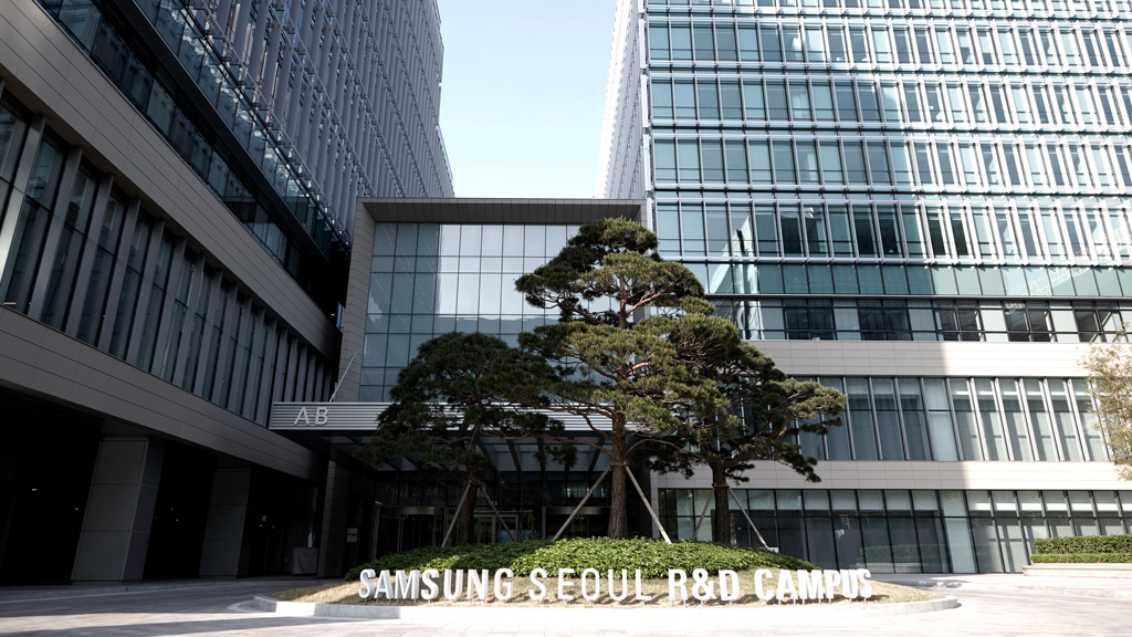 This is an exterior view of the Samsung Seoul R&D Campus. The words “SAMSUNG SEOUL R&D CAMPUS” and  pine trees stand out in front of the building. In the background, you can see a building with a modern glass exterior wall.