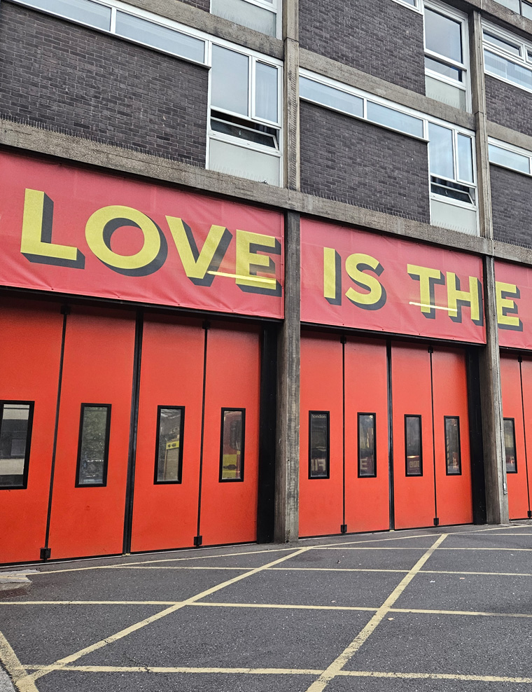 The building facade features bold red doors with the striking phrase 'LOVE IS THE' displayed above, infusing the space with vibrant energy and meaning.