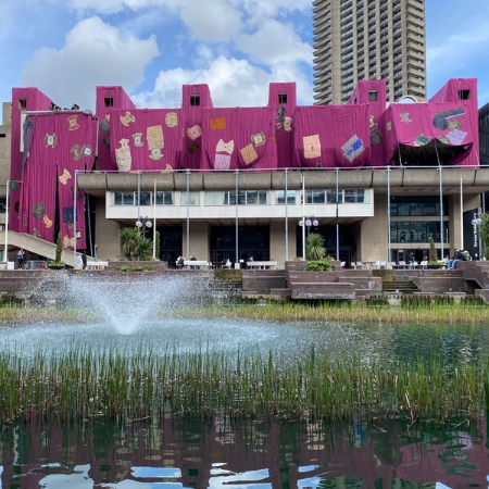 Modern architecture adorned with striking purple drapes and unique decorations catches the eye. The pond and fountain in the foreground add a lively and harmonious touch.