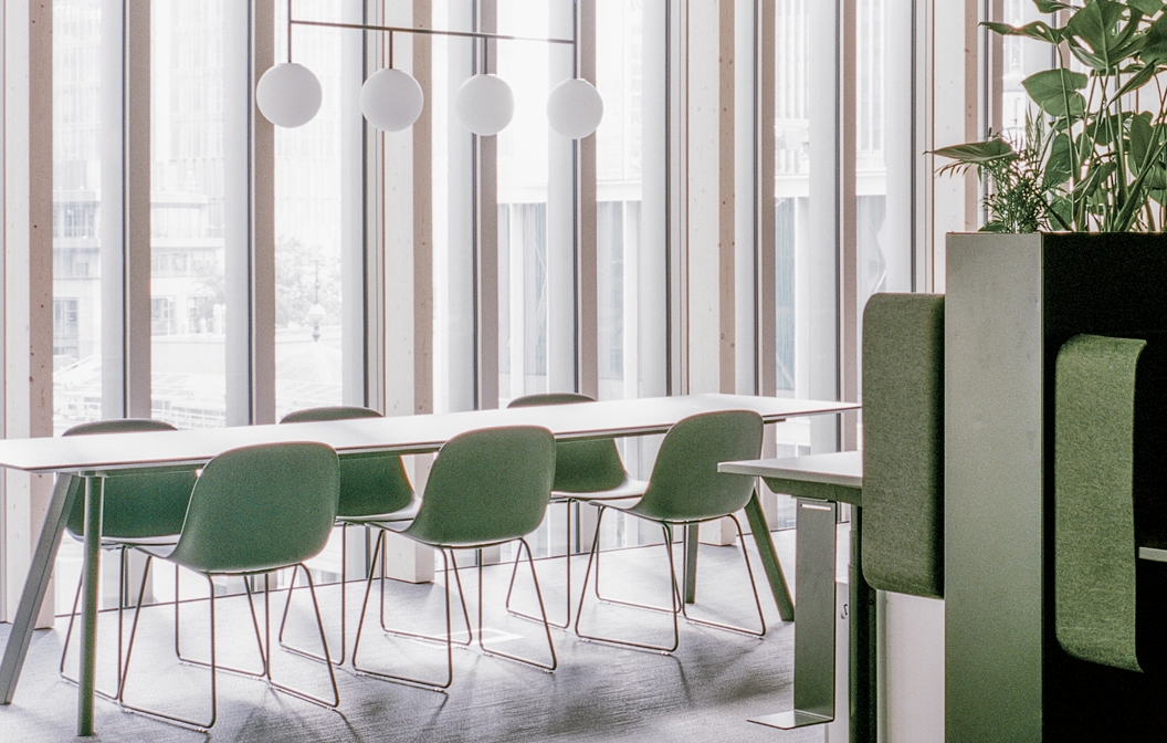 Natural light streaming through large windows illuminates the deep green chairs and minimalist white table, creating a sleek and bright atmosphere.