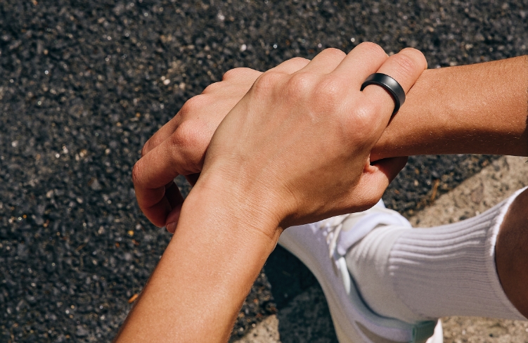 Left hand clasping right wrist. A first-person view from above, showing a Galaxy Ring Titanium Black on the left index finger, white sneakers and socks under the hand, and an asphalt floor in the background.