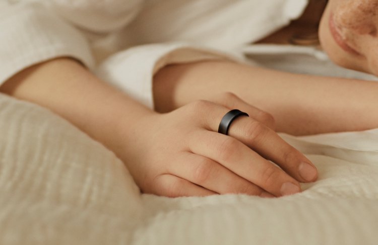  A person is lying on a bed. The person is dressed in white and is wearing a Galaxy Ring Titanium Black on the index finger.