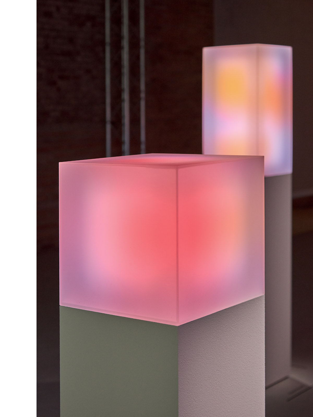 Close-up of a glowing pink cube on a pedestal with a softly lit, colorful cube in the background, part of an art installation with a focus on light and color