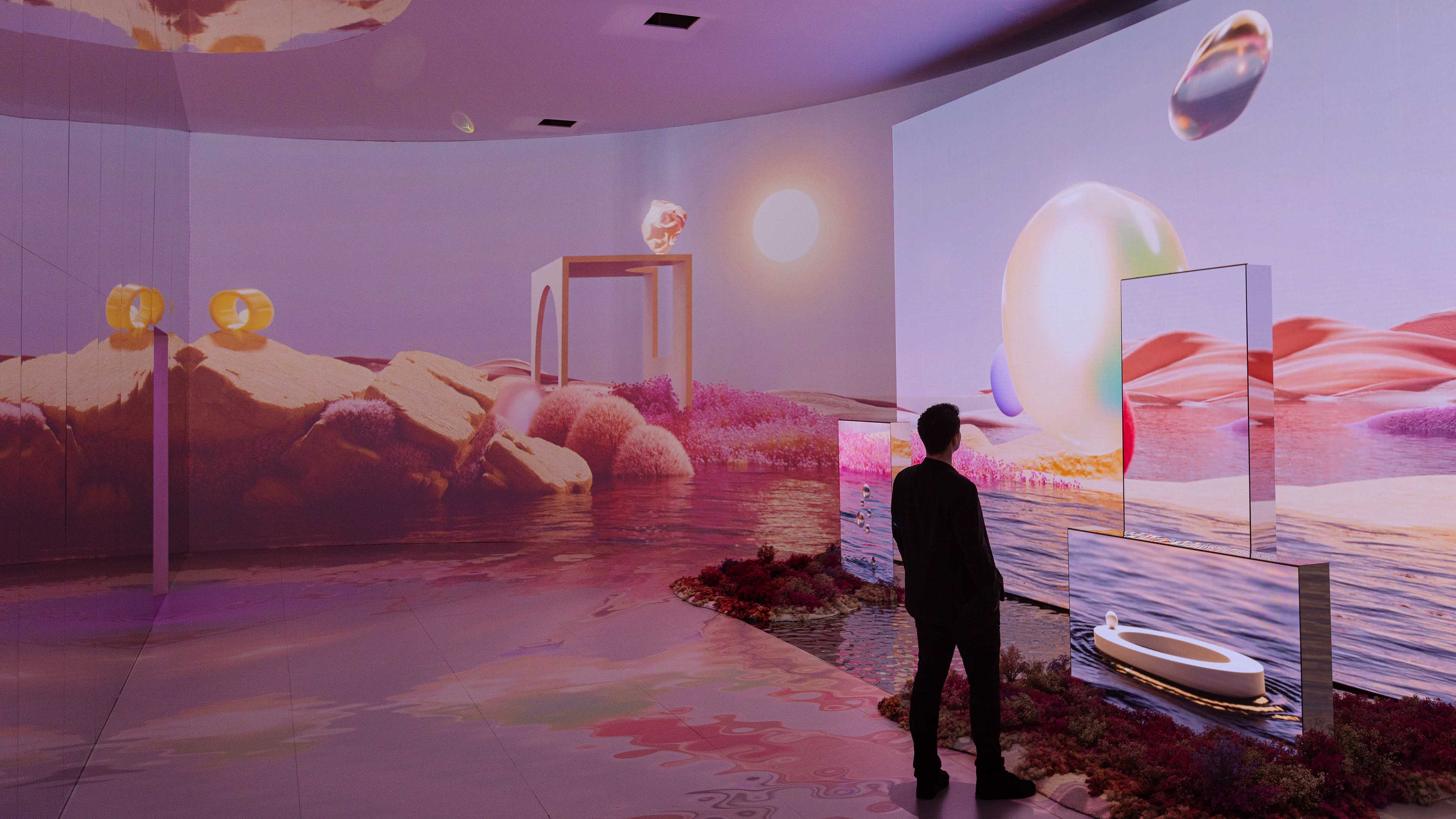 A person stands observing a large-scale, immersive art installation with surreal landscapes projected on walls and panels, creating an otherworldly atmosphere