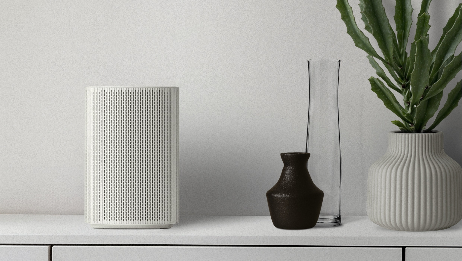 The Premiere 5 device arranged with decorative objects. From left to right on a white cabinet: The Premiere 5, a small brown bottle, a tall clear vase behind it, and a white cactus planter beside them.